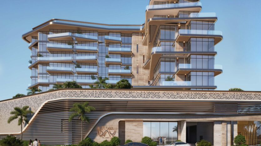A modern multi-story building with curved glass balconies, textured beige walls, and green terraces welcomes you to Flora Bay. Palm trees and parked cars line the entrance under a clear blue sky.