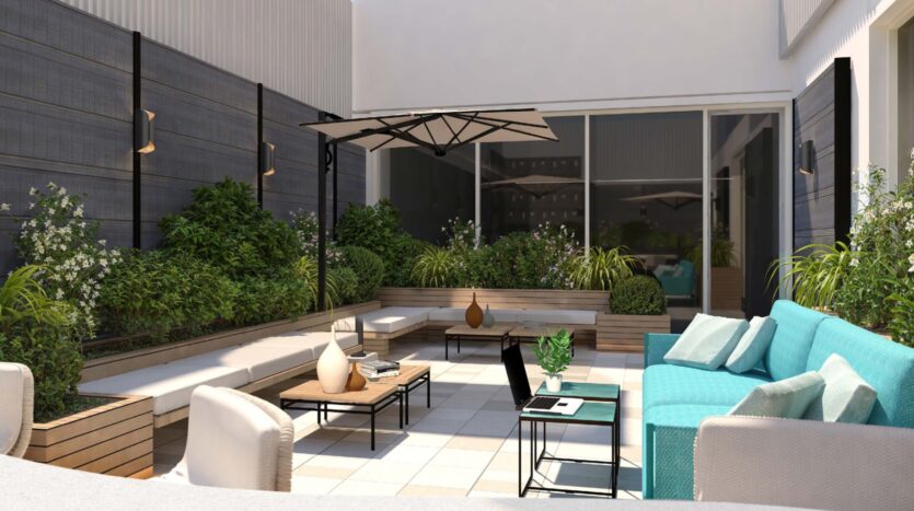 Modern outdoor patio with cushioned seating, coffee tables, potted plants, and a laptop on the table. Flora Bay greenery lines the edges, while glass doors in the background create a comfortable and welcoming atmosphere under a large umbrella.