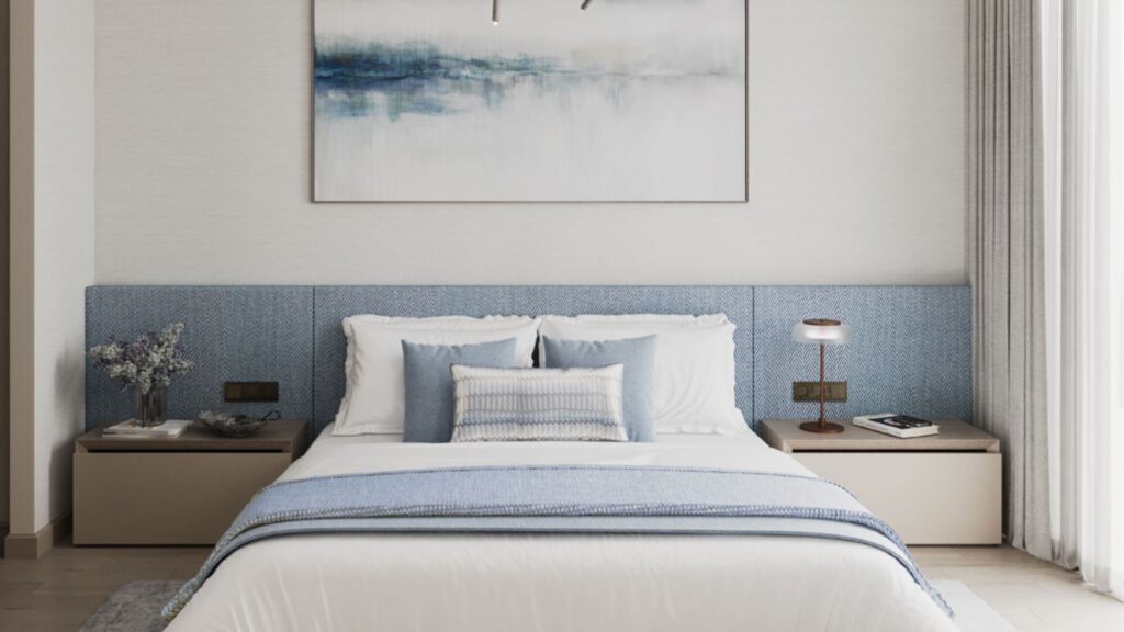 Modern bedroom in the Inara residence with a neatly made bed, blue headboard, white and blue sheets, two beige nightstands with lamps, a vase of flowers, and a large abstract painting above the bed. Natural light comes in from the right.