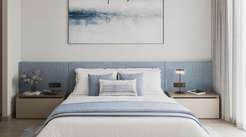 A modern bedroom in the Inara residence features a neatly made bed with white and blue sheets, flanked by beige nightstands. Above the bed hangs an abstract blue and white painting, while a lamp and vase of flowers sit on the nightstands.