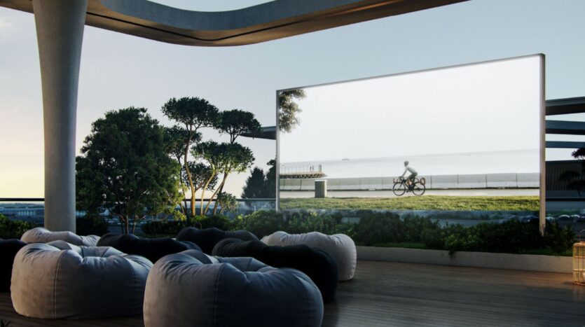 The modern outdoor living room at the Inara residence features beanbag chairs facing a large screen showing a cyclist by the sea, surrounded by greenery and architectural columns under an open sky.