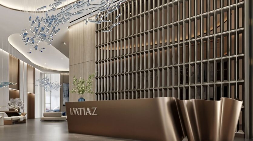 The modern and luxurious lobby of the Inara residence features a sculptural bronze "IMTIAZ" reception desk, artistic pendant lamps, a large decorative wall, and elegant armchairs with contemporary décor.