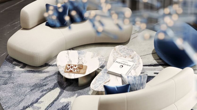 Curved cream sofas with blue pillows surround two marble coffee tables at the Inara residence—one holds desserts and a glass bottle, the other books and headphones—all set on a patterned rug under soft lighting.