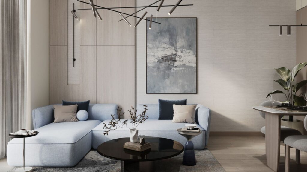 The modern living room of the Inara residence features a light blue sectional sofa, dark throw pillows, a round black coffee table with decorative accents, abstract wall art, minimalist lighting, and a partially visible dining area on the right.