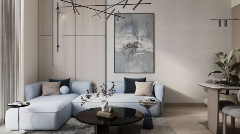 The modern living room of the Inara residence features a light blue sectional sofa, dark throw pillows, a round black coffee table with decorative accents, abstract wall art, minimalist lighting, and a partially visible dining area on the right.