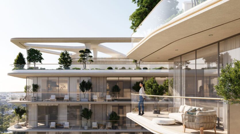 A person stands on a modern balcony at the Inara residence, surrounded by glass railings, plants, and outdoor furniture, overlooking the lush, open terraces of a contemporary multi-story building.