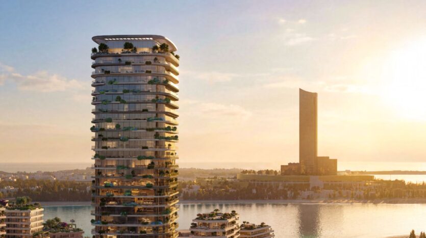 Une tour moderne, Le Château, aux balcons incurvés et à la végétation luxuriante, se dresse au bord de l'eau au coucher du soleil, tandis qu'une autre grande tour s'élève à l'arrière-plan au-dessus des eaux calmes reflétant la chaleur du ciel.