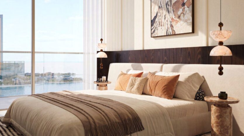 Une chambre à coucher moderne du Château avec un grand lit, des coussins marron et beige et une couverture. Des lampes suspendues, des œuvres d'art murales abstraites, des tables d'appoint et des fenêtres allant du sol au plafond offrent une vue sur l'eau et les bâtiments à l'extérieur.