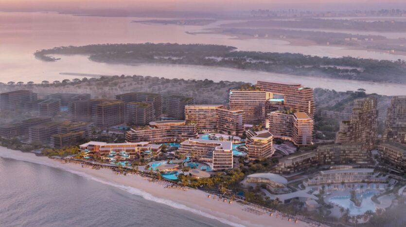 Aerial view of Meriva, a modern seaside resort with numerous terraced buildings, swimming pools, and palm trees along a sandy coastline at sunset, with sparkling water and the city skyline in the background.