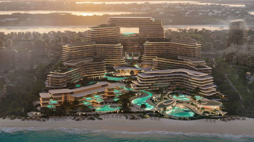 Aerial view of Meriva, a luxury seaside resort featuring modern buildings, large swimming pools, and palm trees with winding paths, set against the backdrop of the ocean at sunset and a distant residential area.