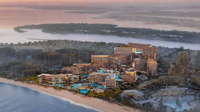Aerial view of the Meriva seaside resort, with its modern terraced buildings, swimming pools, and palm trees along the shore, set against a backdrop of calm waters and distant islands at sunset.