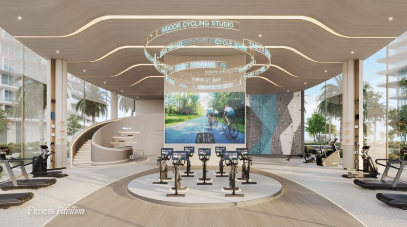 This modern and spacious gym is equipped with Meriva stationary bikes facing a large screen, a curved ceiling bearing the words "Indoor Cycling Studio," treadmills, a climbing wall, and large windows offering a glimpse of the palm trees outside.