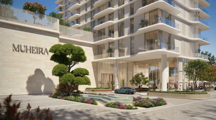 A modern high-rise building with large glass windows bearing the inscription "Muheira." The landscaped area includes trees, plants, a small fountain, and a blue car parked near the Muheira entrance.