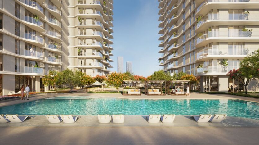 Muheira features a modern residential complex consisting of two large buildings, a large outdoor swimming pool, lounge chairs, and cabanas surrounded by landscaped trees and flowers on a sunny day.