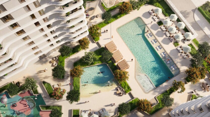 Aerial view of the modern Muheira residential complex with two outdoor pools, lounge chairs, umbrellas, shaded cabanas, lush vegetation, walkways, and a lively playground. People are enjoying the pools and outdoor spaces.