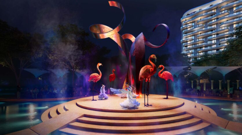 Una scena notturna in Piazza Roma mostra ballerini in costumi bianchi su un palco circolare circondato da grandi sculture di fenicotteri rosa, con un elemento centrale astratto ricurvo e un edificio illuminato sullo sfondo.
