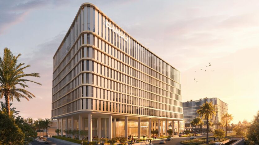 Un moderno edificio a più piani con facciate in vetro si erge al tramonto vicino a Piazza Roma, circondato da palme e viali alberati. Le persone passeggiano all'aperto mentre gli uccelli volano sopra le loro teste. Sullo sfondo è visibile un altro edificio simile.