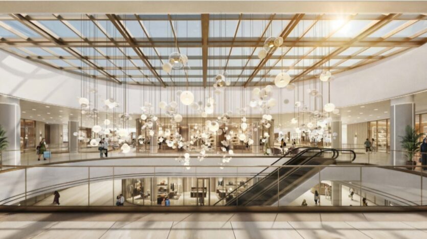 L'interno di un moderno centro commerciale ispirato a Piazza Roma, con un grande soffitto in vetro, luci sospese, scale mobili e negozi su due livelli. La luce del sole entra dalle finestre, creando un'atmosfera luminosa e accogliente per i visitatori.