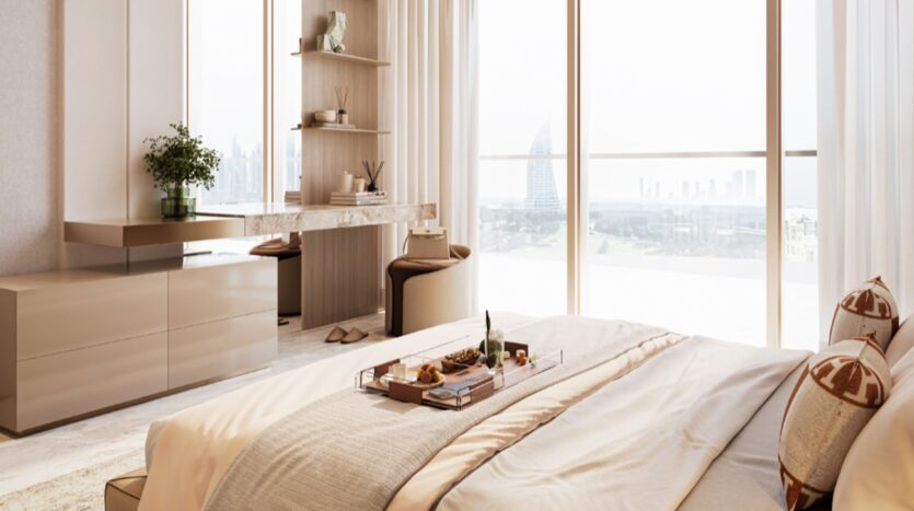 The modern and sunny Sea Cliff room features a large bed with a breakfast tray, a stylish desk, beige tones, floor-to-ceiling windows, and views of the cityscape in the background, including a distinctive sail-shaped building.