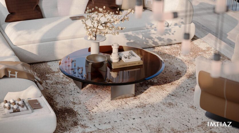 A modern living room in Sea Cliff features a round glass coffee table with books, candles, a bowl, and a vase with branches, surrounded by light-colored sofas on a textured beige rug. The Imtiaz logo appears in the lower right corner.