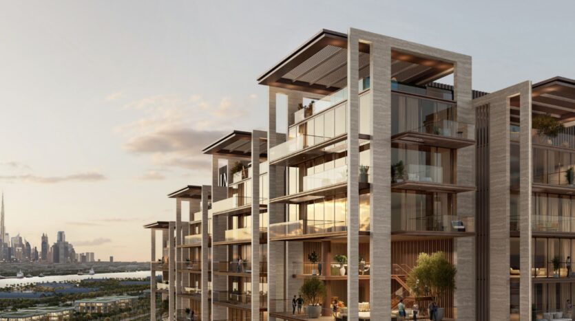 Modern luxury apartment building with large bay windows and spacious balconies, some with greenery. The setting sun reflects off the building's surfaces. In the background, you can see the sea cliffs and the city skyline over the sparkling water.