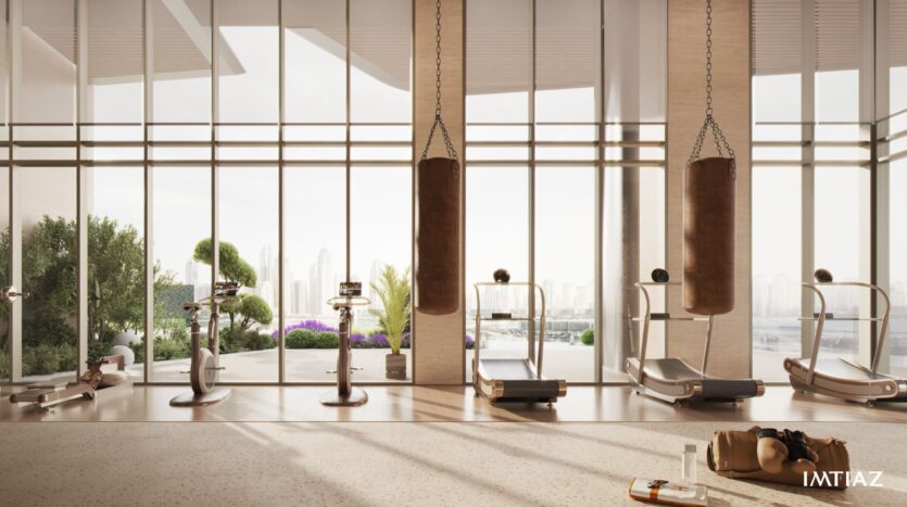 A modern gym with large windows overlooking the Sea Cliff cityscape, featuring treadmills, exercise bikes, punching bags, and a bench with a gym bag and water bottle in the foreground. Lush greenery is visible outside.