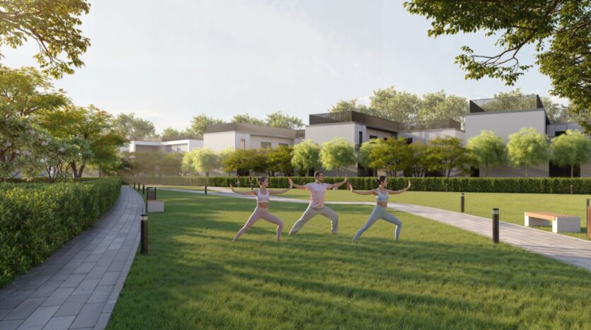 Three people practice yoga on a lawn at Sobha Sanctuary, surrounded by trees, hedges, benches, and contemporary white buildings under a clear sky in a modern residential park.