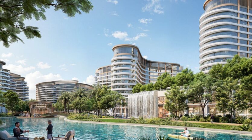 The modern, curved towers of Sobha Sanctuary overlook a landscaped area with palm trees, a large waterfall, and a clear blue pond where people kayak and relax by the water under a bright, sunny sky.