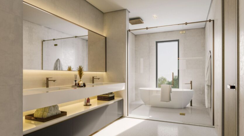 Modern bathroom at Sobha Sanctuary featuring a large double vanity unit, decorative accents, a backlit mirror, a glass bathtub, a towel rack, and a window with natural light. The decor is minimalist with neutral tones and gold accents.