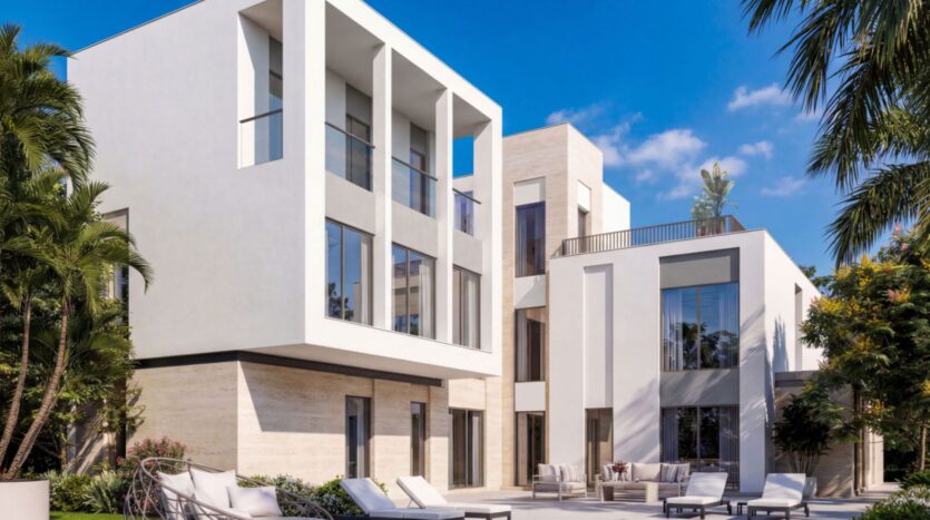 Modern three-story white house in Sobha Sanctuary with large windows, balconies, and an outdoor patio furnished with lounge chairs and a table. Surrounded by palm trees and lush greenery under a bright blue sky.