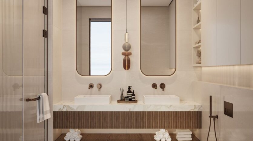 Современная ванная комната в The Residences DIFC с двойной раковиной и прямоугольным зеркалом, мраморной столешницей, свернутыми полотенцами и минималистичными аксессуарами в нейтральной цветовой гамме. Окно обеспечивает естественное освещение, а на полках размещены декоративные предметы.
