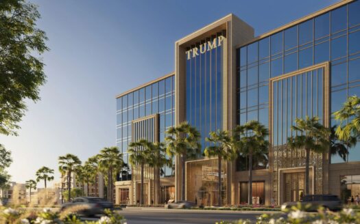 Un edificio moderno con fachada acristalada, palmeras en la parte delantera y la inscripción «TRUMP PLAZA» en la parte superior. Los coches circulan por la calle y el cielo está despejado y azul.
