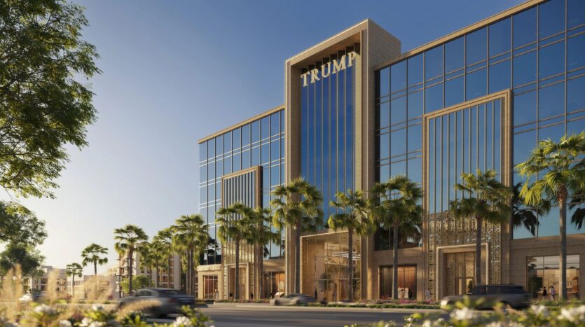 Un bâtiment moderne à la façade vitrée, avec des palmiers à l'avant et la mention "TRUMP PLAZA" au sommet. Des voitures passent dans la rue et le ciel est clair et bleu.
