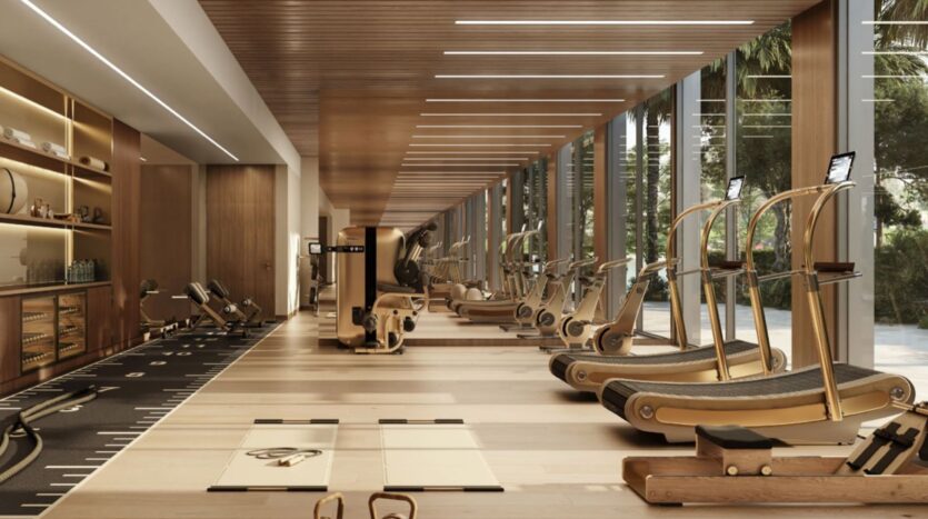 Salle de sport spacieuse et moderne au Trump Plaza avec des sols et des plafonds en bois, de grandes fenêtres, des équipements de fitness dorés, des tapis de course, des poids et des zones d'étirement, avec une lumière naturelle qui pénètre par le côté droit.