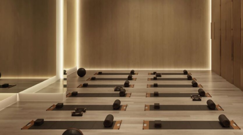 Le studio de yoga moderne du Trump Plaza est doté de sols en bois, de murs neutres et d'un éclairage doux. Des tapis de yoga, des blocs, des sangles et des traversins sont soigneusement disposés en rangées. Un mur en miroir est visible sur le côté gauche.