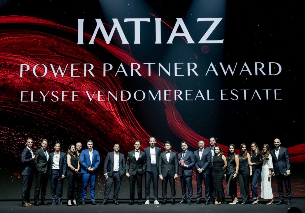 Eine Gruppe von Personen in Abendgarderobe steht auf der Bühne unter einem großen Bildschirm, auf dem „IMTIAZ Power Partner Award Elysee Vendome Real Estate” vor einem spektakulären rot-schwarzen Hintergrund zu sehen ist.