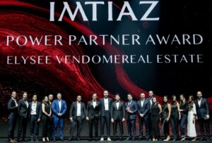 Eine Gruppe von Personen in Abendgarderobe steht auf der Bühne unter einem großen Schild mit der Aufschrift „IMTIAZ POWER PARTNER AWARD ELYSEE VENDOMEREAL ESTATE”, das Dubai Immo repräsentiert, mit einem abstrakten rot-schwarzen Hintergrund hinter ihnen.
