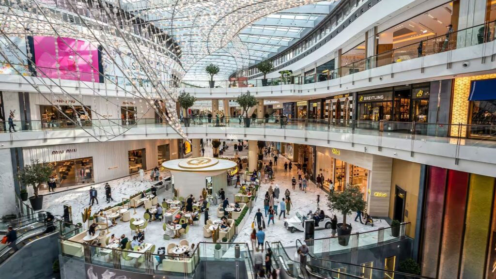 Un centre commercial spacieux et moderne inspiré des grands centres commerciaux de Dubaï, avec plusieurs étages, un plafond de verre, divers magasins et des gens qui profitent d'un espace café central entouré de verdure et d'un éclairage lumineux.