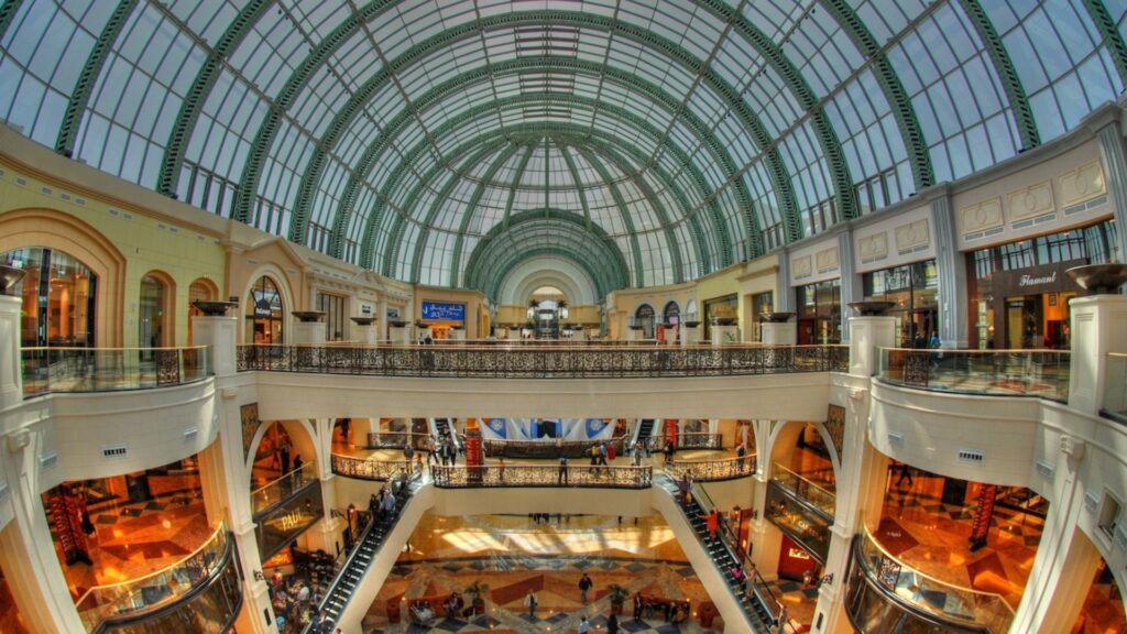 L'intérieur d'un centre commercial spacieux, qui rappelle le Mall of the Emirates, se caractérise par un haut plafond de verre arqué, deux étages de boutiques, d'élégantes rampes et un carrelage à motifs. Les gens se promènent et font leurs achats dans cet espace lumineux.
