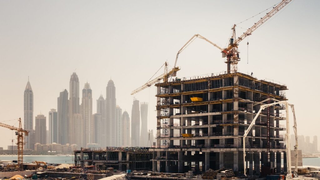 Ein mehrstöckiges Gebäude im Bau mit Kränen im Einsatz, vor dem Hintergrund einer nebligen Skyline aus mehreren großen Wolkenkratzern – ein ehrgeiziges Immobilienprojekt in Dubai mit Materialien und Ausrüstung, die rund um die Baustelle zu sehen sind.