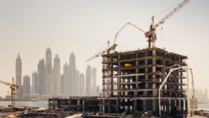Ein mehrstöckiges Gebäude im Bau mit Kränen im Einsatz, vor dem Hintergrund einer nebligen Skyline aus mehreren großen Wolkenkratzern – ein ehrgeiziges Immobilienprojekt in Dubai mit Materialien und Ausrüstung, die rund um die Baustelle zu sehen sind.