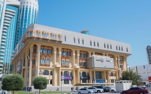 Ein großes Regierungsgebäude mit Bogenfenstern und dekorativen Säulen erhebt sich unter einem strahlend blauen Himmel, umgeben von modernen Wolkenkratzern, die die Dynamik des Immobilienmarktes in Dubai widerspiegeln, Palmen, geparkten Autos und Menschen, die in der Nähe spazieren gehen.