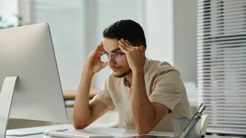 A man wearing glasses is sitting at a desk in front of a computer, holding his head in his hands and looking stressed, perhaps preparing for the Dubai real estate agent exam. The room is very bright and the decor is modern.