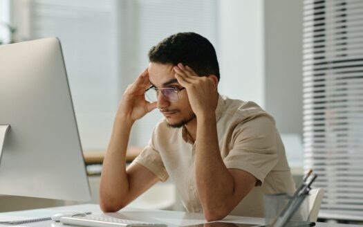 Ein Mann mit Brille sitzt an einem Schreibtisch vor einem Computer, hält seinen Kopf mit beiden Händen und wirkt gestresst, vielleicht bereitet er sich gerade auf die Prüfung zum Immobilienmakler in Dubai vor. Der Raum ist sehr hell und modern eingerichtet.