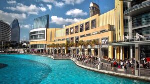 Ein großes, modernes Einkaufszentrum, das mit seinen gelben Glasfassaden an die großen Einkaufszentren in Dubai erinnert, grenzt an einen künstlichen See mit kristallklarem Wasser, während die Menschenmenge unter teilweise bewölktem Himmel die Promenade entlangschlendert.