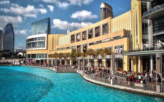 A large modern shopping mall, reminiscent of the large shopping malls in Dubai, with yellow and glass facades, borders a crystal-clear blue artificial lake, while crowds stroll along the waterfront promenade under a partly cloudy sky.