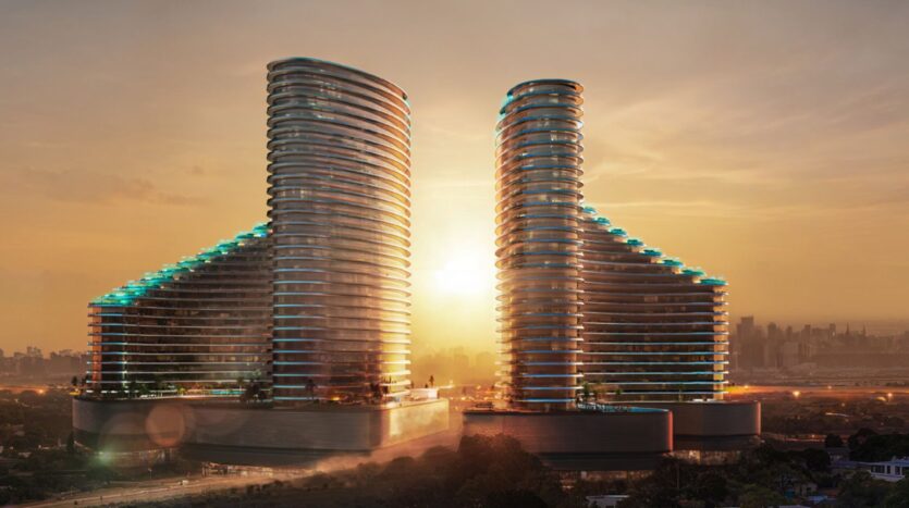 Two modern, curved skyscrapers with illuminated balconies stand side by side at sunset, with the sun shining between them and the city skyline visible in the background.