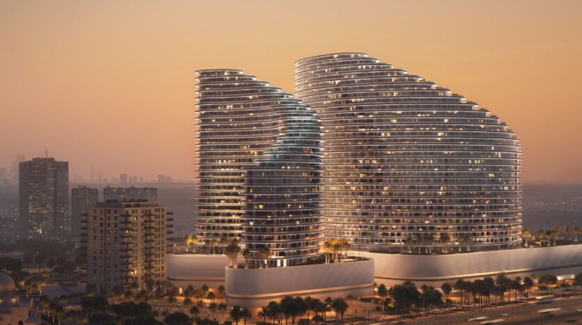 Two modern, curved skyscrapers with illuminated windows stand out against the sunset, their facades glowing like a flame against the cityscape, surrounded by smaller buildings and palm trees against a hazy horizon.