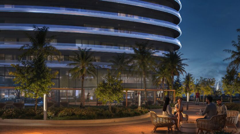 A group of people sit and chat on Skyflame wicker chairs in an outdoor space with palm trees and lights, in front of a modern, curved building at dusk.