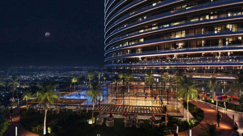 A modern tower, illuminated at night, overlooks the cityscape. The terrace, known as Skyflame, features palm trees, a basketball court, a tennis court, and outdoor seating, all bathed in soft lighting with the moon visible in the sky.